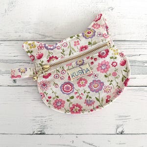 May include: A cat-shaped coin purse made of off-white fabric with a floral pattern in shades of pink, purple, and green. The purse features a gold-toned metal zipper closure and a small rectangular fabric label with the text KUSHA printed on it.
