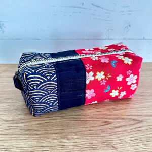 May include: A rectangular zipped pouch with a patchwork design. The pouch features sections of navy blue fabric with a gold wave pattern, solid navy blue, and red fabric with a white and pink floral print. The zip is silver.