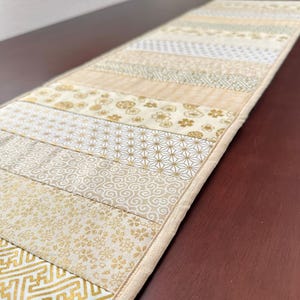 以下が含まれることがあります： パッチワークデザインの長方形テーブルランナー。 ランナーは、金色の花柄と幾何学模様のさまざまなベージュとクリーム色の生地が特徴です。 エッジはクリーム色のボーダーで仕上げられています。