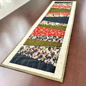 以下が含まれることがあります： パッチワークデザインの長方形テーブルランナー。赤、緑、黒、柄物の生地が交互にストライプ状に配置され、花柄や幾何学模様が施されています。縁は金とクリーム色のボーダーでトリミングされています。