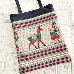 May include: A rectangular tote bag with a woven design featuring a llama and figures in traditional clothing. The bag has a navy blue top and straps. The woven pattern includes red, black, white, and yellow stripes.