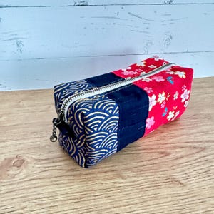 May include: A rectangular pencil case with a zipper closure. The case is made of fabric with three distinct sections: a navy blue section with a gold wave pattern, a navy blue solid section, and a red section with white and pink floral designs.