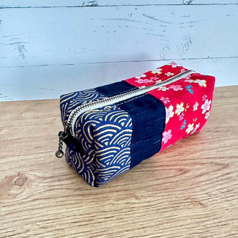 May include: A rectangular pencil case with a zipper closure. The case is made of fabric with three distinct sections: a navy blue section with a gold wave pattern, a navy blue solid section, and a red section with white and pink floral designs.