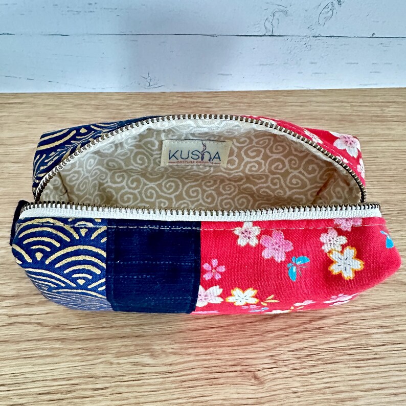 May include: Open pencil case with a zipper, featuring a patchwork design. The case combines navy blue fabric with a wave pattern, solid navy, and red fabric with floral prints. The interior is beige with a swirl pattern, and the brand name "Kusha" is visible.