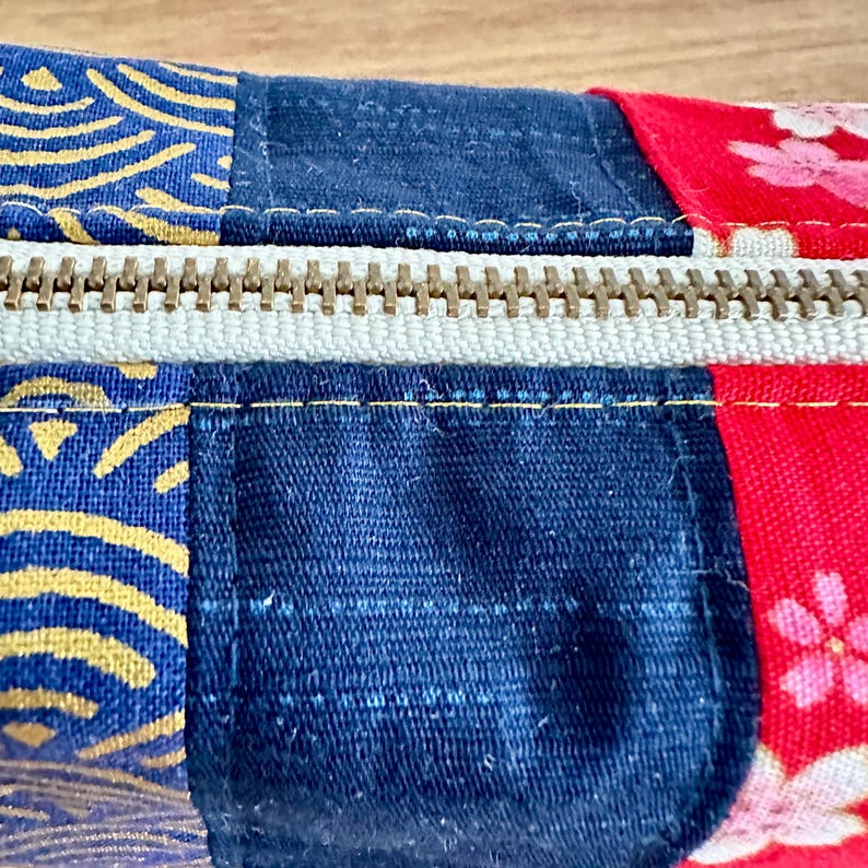 May include: Close-up of a patchwork fabric zipper pouch. The pouch features sections of navy blue, red with floral patterns, and blue with gold wave designs. A beige zipper runs across the top, with visible stitching.