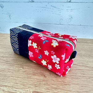 May include: A rectangular pencil case with a zipper closure. The case features three distinct fabric sections: a navy blue section with a gold wave pattern, a solid navy blue section, and a red section adorned with white and pink floral patterns and blue butterflies.