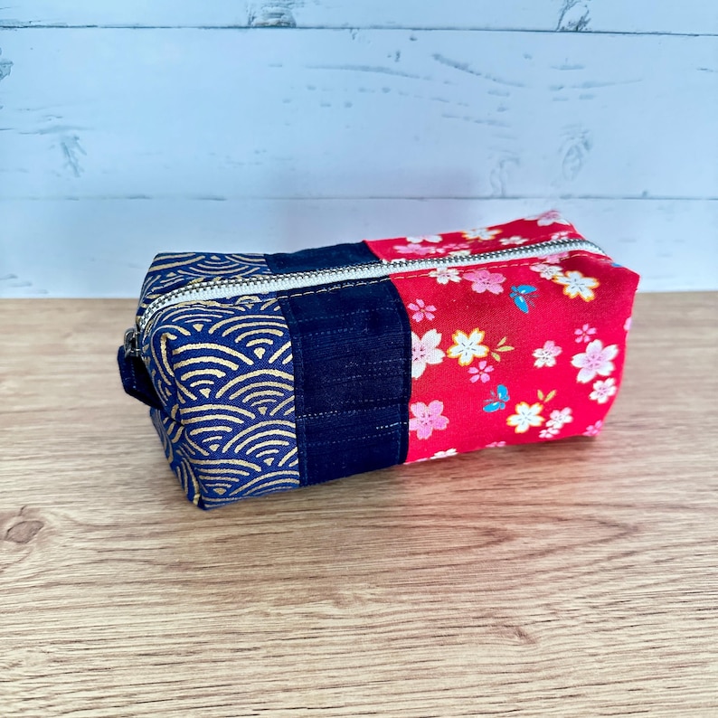 May include: A rectangular fabric pouch with a zipper closure. The pouch is divided into three sections: a navy blue section with a gold wave pattern, a solid navy blue section, and a red section with a floral print. The zipper is silver.