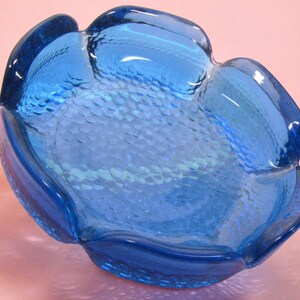 Peut inclure: Un bol en verre bleu vif en forme de fleur à cinq pétales arrondis. La surface intérieure présente un motif texturé en écailles qui capte la lumière, tandis que les bords extérieurs sont lisses et incurvés pour former le design des pétales.
