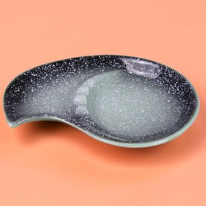 May include: A teardrop-shaped ceramic dish featuring a gradient color scheme that transitions from a dark charcoal black with white speckles on the outer edge to a lighter, muted sage green center with a smooth, glossy finish throughout.