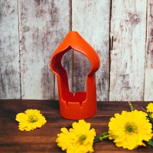 May include: A red ceramic candle holder with a decorative cutout design. The holder is sitting on a wooden surface with yellow flowers.