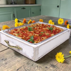 May include: A rectangular white distressed wooden tray with gold-toned metal handles containing a foil pan filled with penne pasta in a red tomato sauce, garnished with fresh green basil leaves, surrounded by scattered yellow and orange daisy flowers.