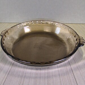 May include: A clear, brown glass pie dish with a scalloped edge and two handles. The pie plate is round and has a slightly raised rim. The dish is made of a durable glass material.