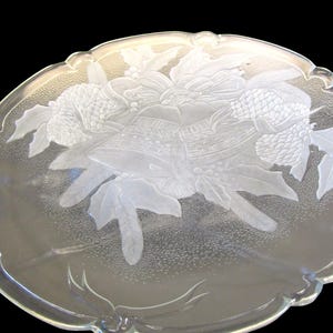 Op de afbeelding: Ovale serveerschaal van helder glas met een geschulpte rand. De schaal heeft een geëtst ontwerp van kerstklokken, dennenappels en bladeren. Het ontwerp is in mat wit, wat een contrast creëert met het heldere glas.