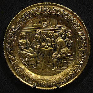 Könnte beinhalten: Eine goldfarbene Zierplatte mit einem detaillierten Relief einer Szene von Menschen, die sich um einen Tisch versammeln. Die Platte hat einen verzierten floralen Rand und einen dunklen Hintergrund.