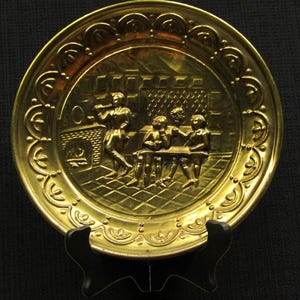 May include: A gold-colored decorative plate with a detailed embossed scene of people gathered around a table. The plate has a decorative border and is displayed on a black stand. The scene includes a fireplace and a person playing a musical instrument.