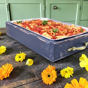 Könnte beinhalten: Ein rechteckiges, dunkelblaues Holztablett mit abgenutzten Kanten und goldfarbenen Metallgriffen, das eine Einweg-Aluminiumschale mit Penne-Nudeln in roter Tomatensoße enthält, garniert mit frischen grünen Basilikumblättern.