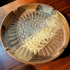 May include: A brown ceramic ashtray with a circular design featuring concentric circles and a raised center. The ashtray has a glazed finish with a light brown and white color scheme.