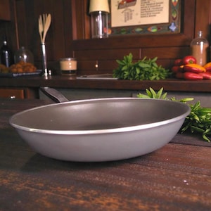 Puede incluir: Una sartén gris antiadherente con borde plateado y mango negro sobre una superficie de madera. Hierbas frescas junto a la sartén. El fondo muestra una cocina con ingredientes y utensilios.