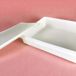 Puede incluir: Un contenedor de almacenamiento rectangular blanco con una tapa a juego, parcialmente abierto. El contenedor está hecho de un material liso y opaco. La tapa es rectangular y descansa junto al contenedor. El fondo es de color rosa liso.