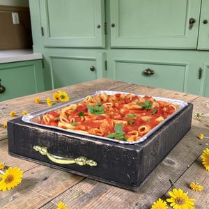 May include: A rectangular black wooden serving tray with a gold-toned metal handle, containing a foil pan filled with penne pasta coated in red tomato sauce and garnished with fresh green basil leaves, surrounded by scattered yellow daisy flowers.
