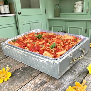 May include: A rectangular gray wooden tray with gold-toned metal handles holds a disposable aluminum foil pan filled with rigatoni pasta in a red tomato sauce, garnished with fresh green basil leaves and scattered yellow flowers.