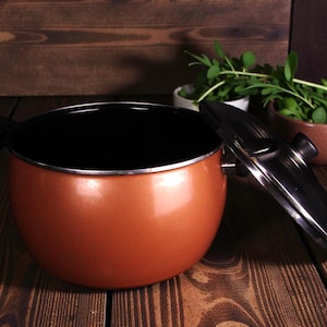 Puede incluir: Una olla de cocina de color cobre con interior negro y tapa a juego. La olla es redonda con un borde plateado y un asa negra. La tapa está parcialmente abierta, revelando el interior. La olla está sobre una superficie de madera.