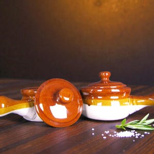 Puede incluir: Tres cuencos pequeños de cerámica para sopa con tapas. Los cuencos son marrones, blancos y amarillos con un asa lateral. Una tapa está quitada. Una ramita de romero y sal están sobre la superficie de madera.