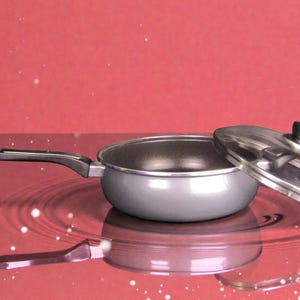 Puede incluir: Una sartén gris con un mango negro y una tapa plateada, sobre un fondo rojo. La tapa está parcialmente abierta, revelando el interior de la sartén. La sartén se refleja en la superficie.