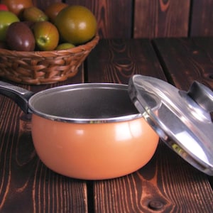 Puede incluir: Una cacerola naranja con un asa negra y una tapa plateada descansa sobre una superficie de madera oscura. Una cesta tejida llena de varias frutas, incluyendo tomates y frutas verdes y marrones, está en el fondo.