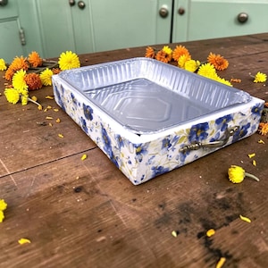 Könnte beinhalten: Ein rechteckiges Serviertablett mit einem Blumenmuster aus blauen und gelben Blüten auf weißem Grund. Das Tablett hat ein metallisch silbernes Inneres und zwei kleine, geschwungene, messingfarbene Metallgriffe an den Seiten.