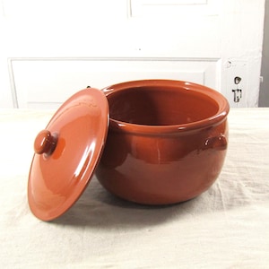 Puede incluir: Una olla redonda de cerámica color terracota con tapa a juego. La tapa está ligeramente entreabierta, revelando el interior de la olla. La olla tiene dos asas pequeñas y está sobre una superficie clara.