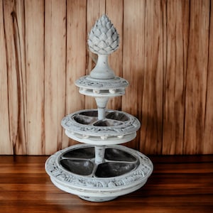 Pode incluir: Um suporte de serviço de madeira branco de três níveis com um remate de abacaxi esculpido. Cada nível tem uma bandeja dividida com um acabamento branco desgastado.