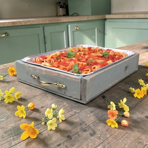 May include: A rectangular gray wooden serving tray with gold-toned metal handles holding a foil pan filled with baked pasta in red tomato sauce, garnished with fresh green basil leaves, surrounded by scattered small yellow and orange flowers.