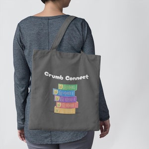May include: A gray tote bag with the text "Crumb Connect" printed in white. A stack of colorful boxes with the text "The First Bread" printed on each box is centered on the bag.