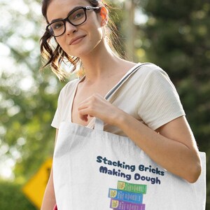 Op de afbeelding: Een witte tote bag met blauwe tekst die "Stacking Bricks Making Dough" zegt. De tas heeft een stapel kleurrijke speelblokken met de tekst "Making Dough" op elk blok.