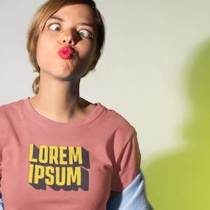 Puede incluir: Una camiseta rosa polvoriento con el texto "LOREM IPSUM" en amarillo, contorneado en negro. La persona lleva una sudadera con capucha azul claro y vaqueros negros. El fondo es una mezcla de blanco y verde lima.