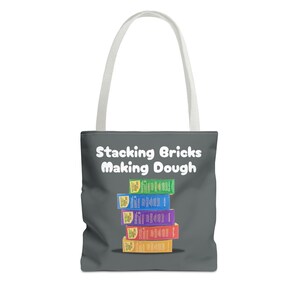 May include: A gray tote bag with white handles featuring the text "Stacking Bricks Making Dough." The bag displays a stack of colorful boxes, each with a different color and design. The boxes are stacked on top of each other.