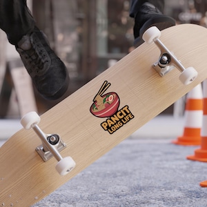 May include: A skateboard with a sticker featuring a bowl of noodles with chopsticks. The sticker has the text "PANCIT LONG LIFE" in a yellow and black font. The skateboard is light brown with silver hardware and white wheels.