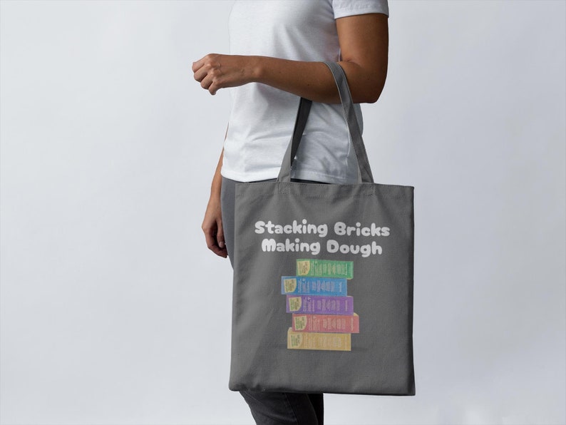 May include: A gray tote bag with the text "Stacking Bricks Making Dough" in white. The bag features a graphic of a stack of colorful toy blocks. The blocks are in shades of blue, green, purple, and yellow.