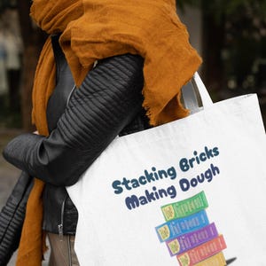 Op de afbeelding: Een witte tote bag met de tekst "Stacking Bricks Making Dough" in blauwe letters. De tas heeft een stapel kleurrijke dozen met dezelfde tekst.