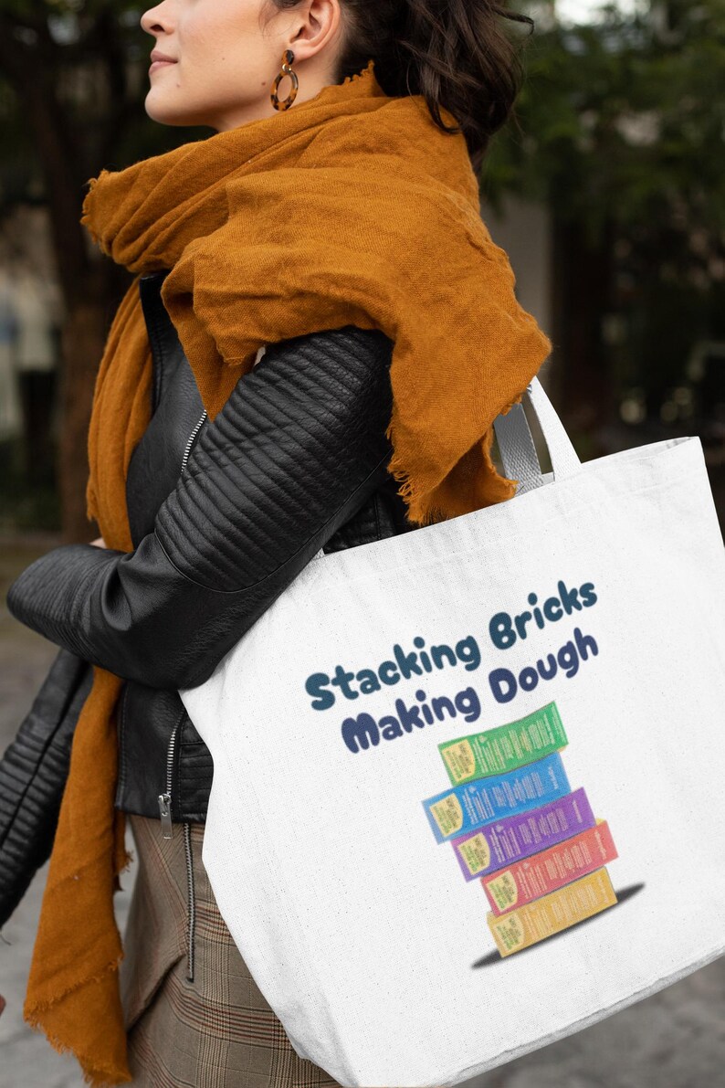 Op de afbeelding: Een witte tote bag met de tekst "Stacking Bricks Making Dough" in blauwe letters. De tas heeft een stapel kleurrijke dozen met dezelfde tekst.