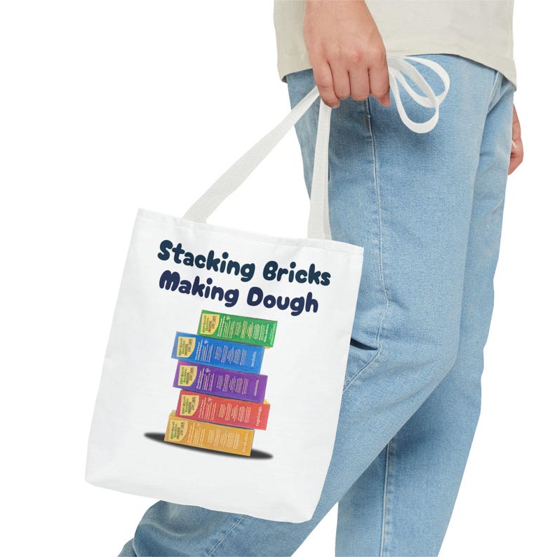 Op de afbeelding: Witte tote bag met de tekst "Stacking Bricks Making Dough" in blauw. Een stapel kleurrijke dozen met de tekst "Play-Doh" staat op de tas.