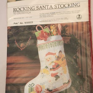 以下が含まれることがあります： 透明なプラスチックパッケージに入ったBernat Rocking Santa Stockingクロスステッチキット。ラベルにはRocking Santa Stocking to Crosstitch、Pak No. W00023と記載されており、約30.48cm x 40.64cmのサイズが示されています。画像には木馬に乗ったサンタクロースが描かれています。