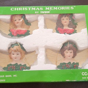Puede incluir: Un juego de cuatro adornos de ángel Christmas Memories by Mann en una caja verde. Cada adorno presenta una cabeza de ángel esculpida con alas, hojas de acebo y bayas rojas. La caja incluye el texto Christmas Memories by Mann y CC-85.