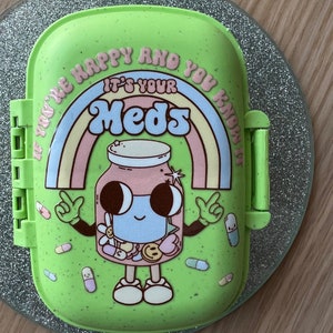 May include: A light green rectangular pill case with a speckled finish. The lid features a graphic of a smiling jar filled with pills, a rainbow, and the text "IF YOU'RE HAPPY AND YOU KNOW IT IT'S YOUR MEDS" printed in a curved, colourful font.