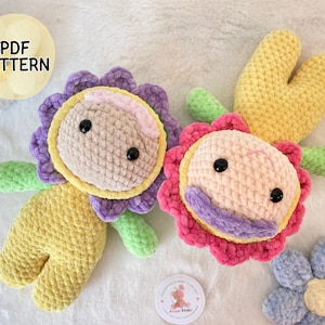 May include: Two crocheted flower-shaped plushies. One is yellow with a purple flower and green leaves. The other is pink with a purple flower and green leaves. Both have black eyes and a pink nose.