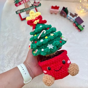 May include: A crocheted Christmas tree ornament. The green tree is decorated with festive embellishments and a candy cane. It's planted in a red pot with a smiling face and brown pom-pom feet. A yellow star and red bow crown the tree.