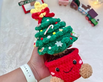 Puede incluir: Adorno navideño hecho a mano. El árbol verde está adornado con adornos y un bastón de caramelo. Está plantado en una maceta roja con una cara sonriente y pies de pompón marrón. Una estrella amarilla y un lazo rojo coronan el árbol.