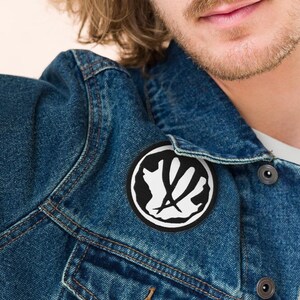 May include: A man wearing a blue denim jacket with a black and white hand patch on the left side of the chest.