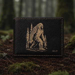 May include: Black bi-fold wallet with a Bigfoot design in gold. The wallet features a textured surface and gold stitching around the edges. The Bigfoot graphic is centered, with trees on either side. The initials "RLV" are in the bottom right corner.